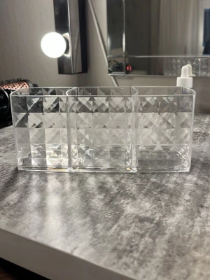 Transparent sminkorganiserare i plast - Snygg och praktisk transparent sminkorganiserare i plast med tre fack. Perfekt för att hålla ordning på borstar, pennor eller andra skönhetsverktyg. Rutigt mönster på sidorna ger en modern känsla och passar bra på sminkbordet.