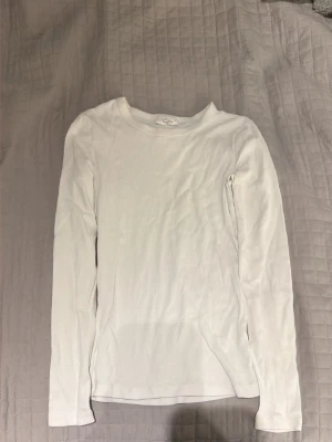 Vit långärmad topp från Gina Tricot - Klassisk vit långärmad topp från Gina Tricot, kollektionen Basically Basics. Tillverkad i ribbad bomull med rund halsringning och figurnära passform. Perfekt att styla lager på lager eller bära som den är.