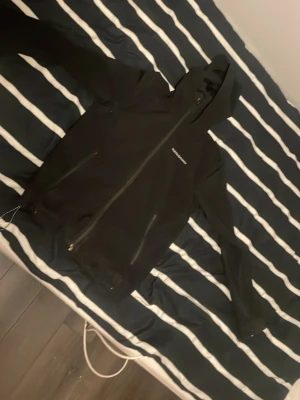 Svart vindjacka från Peak Performance - Svart vindjacka med huva från Peak Performance. Jackan har hel dragkedja framtill, två sidofickor med dragkedja och diskret logga på bröstet. Materialet är slätt och vattenavvisande, perfekt för blåsiga dagar.