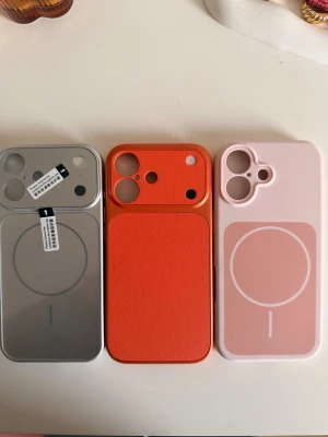 Tre iphone 17 mobilskal MagSafe i olika färger - Säljer tre snygga mobilskal med MagSafe-kompatibilitet. Ett i silver med spegelyta, ett i orange med läderstruktur och ett i ljusrosa med vit MagSafe-ring. Alla har öppningar för kamera och knappar, perfekta för dig som vill ha stil och funktion.