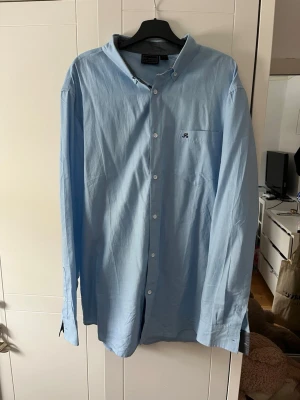 Ljusblå skjorta  - Klassisk ljusblå skjorta med button-down krage och diskret logga broderad på bröstet. Skjortan har långa ärmar och knäppning framtill. Perfekt till jeans eller chinos för en clean och stilren look.