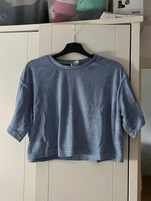 Ljusblå croppad topp från H&M - En ljusblå croppad topp från H&M med korta ärmar och rund halsringning. Materialet är frotté vilket ger en mjuk och skön känsla. Perfekt för dig som gillar en avslappnad och trendig stil.