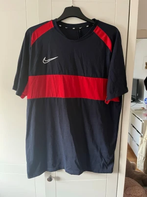 Nike marinblå t-shirt med röd detalj - Snygg marinblå t-shirt från Nike med en bred röd rand över bröstet och röda axelpartier. T-shirten har en vit Nike-logga på vänster sida och är tillverkad i mjukt bomullsmaterial. Perfekt för en sportig och avslappnad stil.