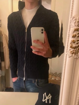 Mörkblå kabelstickad ziptröja - Snygg mörkblå kabelstickad tröja är med metallycklås framtill och hög krage. Tröjan har ribbade muddar vid ärmslut och nederkant höjer sopranstilen. Perfekt till jeans och t-shirt för en clean look.