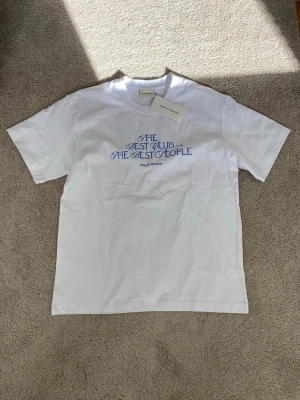 Vit t-shirt från Drôle de Monsieur - Vit t-shirt från Drôle de Monsieur med blå text på bröstet där det står 'The best club for the best people'. Klassisk passform och rund halsringning. Perfekt för dig som gillar stilrena plagg med en twist.