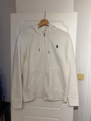 Polo kofta  - Säljer en stilren vit hoodie från Polo Ralph Lauren med dragkedja framtill och klassisk huva med dragsko. Broderad logga i marinblått på bröstet och två fickor framtill. Perfekt för en clean och avslappnad look.