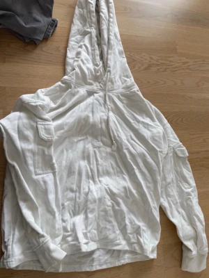 Vit hoodie med fickor och huva - Säljer en vit hoodie med stor huva och dragsko. Hoodien har fickor med lock på båda ärmarna och ribbade muddar vid ärmsluten. Tillverkad i mjukt bomullstyg, perfekt för en avslappnad stil.