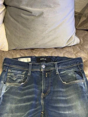 Replay Anbass jeans blå denim - Snygga Replay Anbass jeans i klassisk blå tvätt med slitningar och lätt distressed detalj på ena benet. Modellen har fem fickor, kontrastsömmar och normal passform. Nypris 1499 mitt pris 499