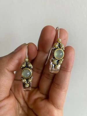 Unika silverörhängen med pärldetaljer - Säljer ett par handgjorda örhängen i silver med guldfärgade detaljer och små vita pärlor. Örhängena har en rund, ljusgrön sten i mitten och dekorativa pärlor runt om. Hängande modell med snygg vintagekänsla och cool design.material is copper