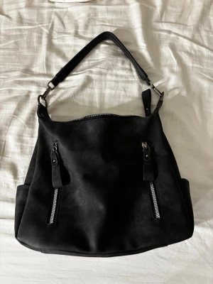 Svart handväska med dragkedjor - Stilren svart handväska i slätt konstläder med två dragkedjefickor framtill och en extra ficka på baksidan. Väskan har en bred axelrem i matchande material och silverfärgade metalldetaljer. Perfekt för dig som vill ha en rymlig och snygg väska till vardagen. Finns en längre axelrem till också