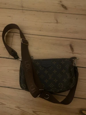 Brun axelväska - Snygg axelväska med klassiskt brunt monogrammönster och bred, justerbar axelrem i mörkbrunt tyg med logotyp. Väskan har silverfärgade metalldetaljer och dragkedja upptill. Perfekt för dig som gillar lyxig och tidlös design.