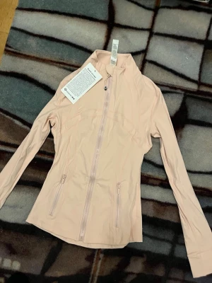 Ljusrosa lululemon zip tröja  - Snygg ljusrosa tröja  från Lululemon med figurnära passform. Tröjan har dragkedja framtill, två fickor med dragkedja och hög krage. Tillverkad i ett mjukt syntetmaterial som passar perfekt till träning eller vardagsoutfits.