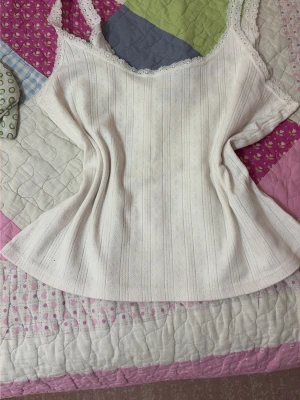 Vit spetsdetaljerad linnetopp - Söt vit linnetopp med tunna axelband och spetsdetaljer längs ringningen. Toppen har ett diskret hålmönster i vertikala ränder och är tillverkad i ett mjukt, lätt material som passar perfekt till sommaren.