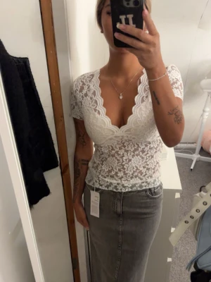 Vit spetsblus med v-ringning - Säljer en superfin vit blus i spets med djup v-ringning och korta ärmar. Toppen har ett blommigt spetsmönster och är croppad med vågig kant nedtill. Perfekt för dig som vill ha en romantisk och trendig look.