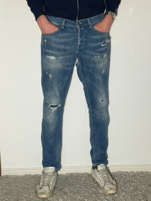 Blå dondup jeans med schyssta slitningar skinny fit - Säljer ett par blå jeans med smal passform och slitna detaljer samt hål på benen. Jeansen har klassisk femficksmodell och knappgylf. Perfekta för en avslappnad och trendig look. 