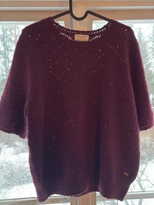 Vinröd stickad topp  - Mysig vinröd stickad topp från Holly & White med korta ärmar och rund halsringning. Tröjan har en luftig stickning och en liten metallbricka nere vid fållen. Perfekt för lager på lager och en trendig look. Helt oanvänd