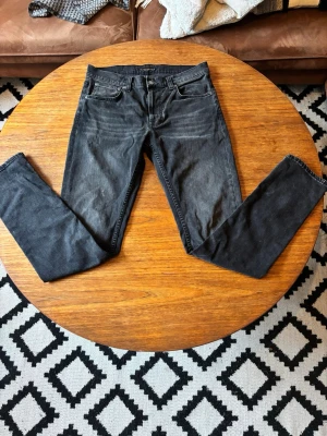 Svarta nudie jeans - Snygga svarta slim fit jeans med klassisk femficksdesign och dragkedjegylf. Jeansen har en något tvättad look och raka ben. Perfekta för dig som gillar en stilren och enkel vibe. Materialet är bomullsdenim med lite stretch för extra komfort.