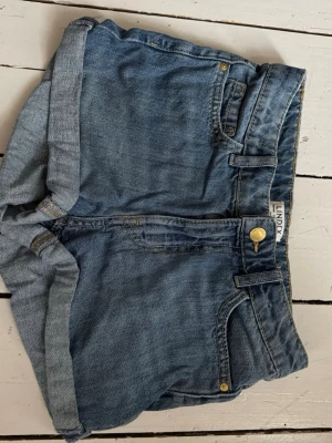 Blå jeansshorts från Lindex - Snygga blå jeansshorts från Lindex med klassisk femficksdesign och uppvikta benslut. Shortsen har gylf med dragkedja och knapp, samt bälteshällor. Perfekta för varma dagar och enkel att matcha med allt.