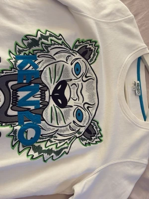 Vit sweatshirt med tiger från Kenzo - Säljer en vit sweatshirt från Kenzo med det ikoniska tigermotivet broderat i blått, grönt och svart på bröstet. Har använt fåtal gånger. Tröjan har rund halsringning och är tillverkad i mjukt bomullsmaterial. Perfekt statement-plagg för dig som gillar streetwear och snygga detaljer. Tre jättesmå fläckar som knappt syns som man sänert kan få bort i tvätten.