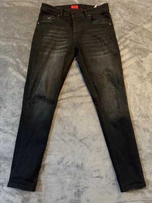 Svarta  jeans från Hugo Boss - Snygga svarta jeans från Hugo Boss med diskreta slitningar framtill och klassiska fem fickor. Jeansen har en smal passform och stretchigt material för extra komfort. Perfekta för dig som gillar en stilren och modern look. 