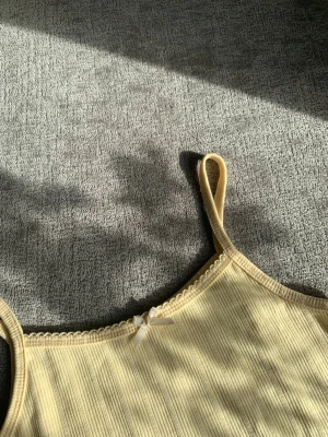 Ljusgul pointelle linne med rosett - Supersöt linnetopp i pointelle med smala axelband och en liten rosett framtill. Perfekt för varma sommar dagar och enkel att styla till olika outfits. Mjuk och bekväm känsla.