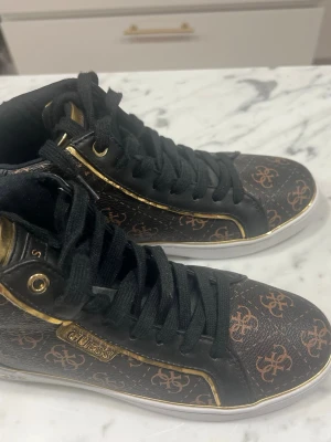 Svarta Guess sneakers med guldiga detaljer - Snygga svarta sneakers från Guess med bruna monogram och guldiga detaljer. Skorna har svarta skosnören, vita sulor och Guess-logga på sidan. Materialet är konstläder med metallicinslag och klassisk high-top design.
