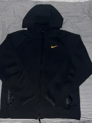 Nocta tech fleece - Snygg svart hoodie från Nike med dragkedja och huva. På bröstet sitter en gul Nike Swoosh som ger en sportig vibe. Hoodien har två fickor med dragkedja och är tillverkad i mjukt material som känns skönt mot huden. Perfekt för dig som gillar streetwear och sportig stil.