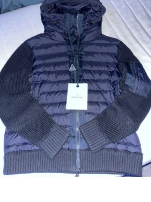 Mörkblå pufferjacka från Moncler - Snygg mörkblå pufferjacka från Moncler med stickade ärmar och muddar. Jackan har huva, dragkedja framtill och Moncler-logga på ärmen. Materialmix av quiltad nylon och stickat tyg ger en modern look. Perfekt för kalla dagar.