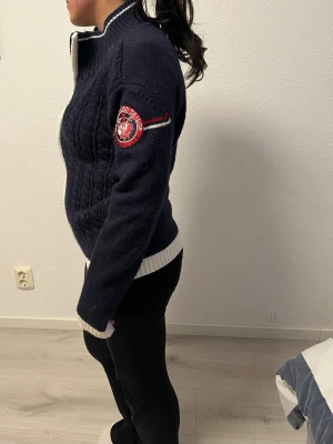 Marinblå stickad tröja från polo edition - Mörkblå stickad tröja från Napapijri med hög krage och dragkedja. Tröjan har vita och röda detaljer, ribbstickade muddar och en tydlig logga på ärmen. Perfekt för kalla dagar och har en sportig, klassisk look. 