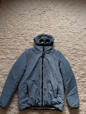 Grå vindjacka med huva och glasögon från CP company - Snygg grå vindjacka med hel dragkedja framtill och två fickor med dragkedja. Jackan har en unik huva med inbyggda glasögon,  Tillverkad i lätt och vindtätt material.