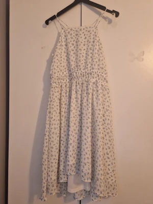 Vit blommig sommarklänning - Söt vit klänning med små blommor i blått, smala axelband och avslappnad passform. Klänningen har resår i midjan och är tillverkad i ett lätt, luftigt material som passar perfekt för varma dagar.Det är dubbeltyg så att man inte ser igenom. 