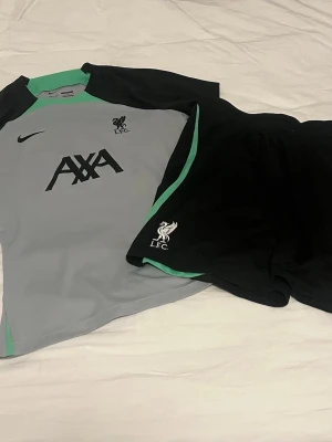Liverpool träningsset Nike grå/svart - Snyggt träningsset från Nike och Liverpool FC med grå tröja och svarta shorts. Tröjan har svarta och mintgröna detaljer samt AXA-logga och LFC-märke. Shortsen är svarta med mintgrön kant och LFC-logga. Perfekt för fotbollsträning eller gym. Köpt utanför Anfield i Liverpool shopen 