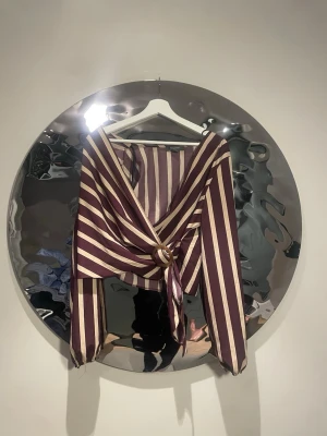 Randig omlottblus med ringdetalj - Snygg omlottblus med vinröd och beige vertikala ränder. Blusen har långa ärmar och en dekorativ ringdetalj i fornt vid midjan. Djup v-ringning och knytband framtill ger en trendig look. Perfekt att styla med hö success eller jeans.