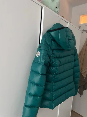  dunjacka från Monclear - Säljer en grön dunjacka från Moncler med huva och svarta detaljer. Jackan har två dragkedjeförsedda fickor framtill, quiltad design och Moncler-logga på ärmen. Perfekt för lite varmare vinterdagar och riktigt snygg till streetwear-stilen.