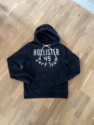 Svart Hollister hoodie med tryck - Svart hoodie från Hollister med vit text 'Hollister 49 Surf Team' på bröstet. 