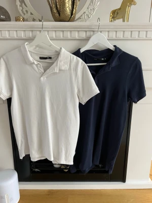 2-pack pikétröjor i vitt och marinblått - Två klassiska pikétröjor från lager 157, en i vitt och en i marinblått. Båda har krage, kort ärm och knappar framtill. Perfekta för en clean och sportig look. Tillverkade i mjuk bomull för skön känsla hela dagen.