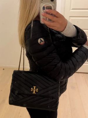 Svart axelväska från Tory Burch - Snygg svart axelväska från Tory Burch med quiltat mönster och guldfärgad kedjerem. Väskan har en elegant guldig logga framtill och är tillverkad i läder. Perfekt för dig som vill ha en stilren och trendig accessoar. Jag köpte den för ca 1år sedan för 8000kr i butik, inga alls defekter och är helt som ny. ÄLSKAR denna väska men har för många!✨