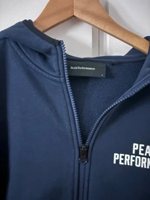 Mörkblå hoodie Peak Performance - Mörkblå hoodie från Peak Performance med dragkedja framtill, huva och två fickor. Klassisk logga i vitt på bröstet. Mjuk insida och ribbade muddar vid ärmslut och nederkant. Perfekt för en avslappnad och sportig stil.
