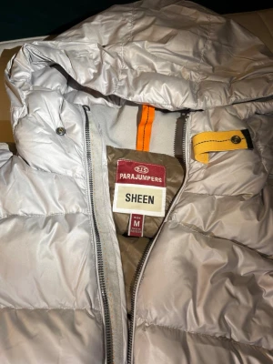 Parajumpers pharell sheen jacka - Snygg silverfärgad dunjacka från Parajumpers med huva, två sidofickor med dragkedja och klassisk patch på ärmen. Jackan har quiltad design och detaljer i orange och gul vid halsen. Perfekt för kalla vinterdagar.M passar S. Kvitto med följer inköpt för 6200kr då fick den som julgåva 