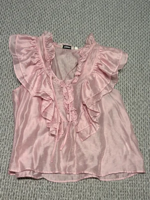Rosa volangblus  - Supersöt ljusrosa blus i lite ”glansigt” material. Blusen har stora volanger längs ringningen och axlarna samt knytband framtill. Ärmlös modell som ger en romantisk och trendig look. Perfekt att styla med jeans eller kjol för en chic vibe. Blusen har en luftig modell med tunnt tyg, vilket passar perfekt för lite varmare dagar!🌸🩰