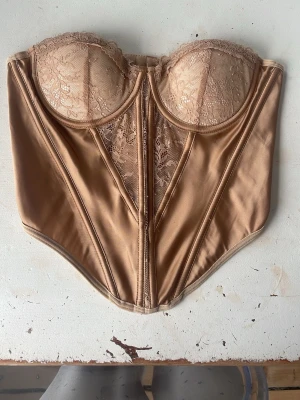 Beige spetskorsett med bygel strl S - Snygg beige korsett med bygel och vadderade kupor. Framsidan har dekorativa spetsdetaljer och slät satinliknande yta. Baksidan har hyskknäppning och spetsinslag. Perfekt för att skapa en feminin siluett och ge stöd. Materialet är en mix av spets och satin.