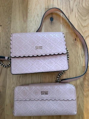 Ljusrosa handväska från Guess - Söt ljusrosa handväska från Guess med matchande plånbok. Väskan har kedjedetaljer på axelremmen och präglat logomönster över hela ytan. Flärpen har vågig kant och Guess-loggan i guld framtill. Perfekt för dig som gillar stilrena accessoarer. Väskan är i gott skick och plånboken har slitage och är alltså i mindre gott skick 