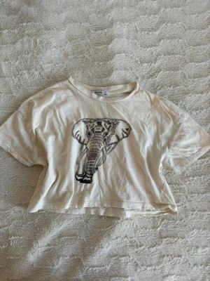 Krämvit croppad t-shirt med tryck på - Snygg croppad t-shirt med ett coolt elefanttryck framtill. T-shirten har korta ärmar och är gjord i mjuk bomull, perfekt för en avslappnad och trendig look. I storlek XS och fins en defekt på sista bilden.