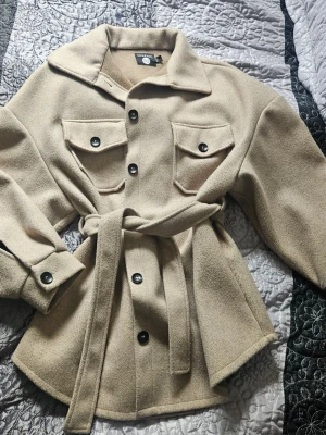 Beige kappa med bälte och fickor - Stilren beige kappa från Loavies med knappar framtill, två bröstfickor och ett matchande midjebälte. Kappan har bred krage, långa ärmar och är tillverkad i ett mjukt, ull-liknande material. Perfekt för lager på lager och trendig look.