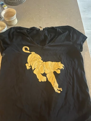 Svart t-shirt med guldtigertryck - Cool svart t-shirt med v-ringning och ett stort guldigt tigertryck på bröstet. Perfekt för dig som vill sticka ut med en unik och trendig look. T-shirten har korta ärmar och är tillverkad i mjuk bomull.