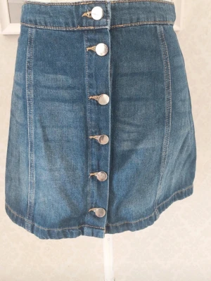 Blå jeanskjol med knappar framtill - Snygg blå jeanskjol i kort modell med silverfärgade knappar längs hela framsidan. Kjolen har klassiska sömmar och en rak passform som ger en tidlös denimlook. 