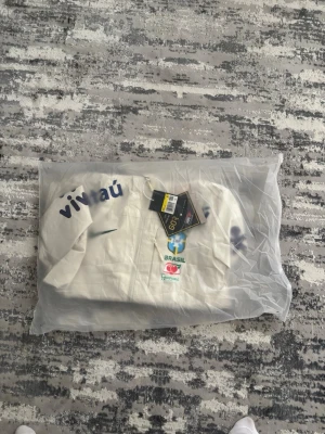 Vit Brasil Nike vindjacka med huva - Snygg vit vindjacka från Nike med Brasil-logga och sponsormärken på bröstet.  Obs , prislappen är ej kvar . Bilderna är tagna för 2 veckor sen men jackan är använd en gång som test ! 