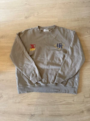 Beige sweatshirt med brodyr från woodbird - Snygg beige sweatshirt med broderade detaljer på bröstet, en röd och gul logga samt en blå och gul text med stjärnor. Rund halsringning och ribbade muddar vid ärmslut och nederkant. Perfekt för en avslappnad stil.