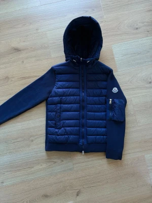 Mörkblå Cardigan - Snygg mörkblå pufferjacka från Moncler med huva och dragkedja framtill. Jackan har quiltad framsida och ärmar i stickad kvalitet samt en praktisk ficka med dragkedja på ena ärmen. Perfekt för kalla dagar och riktigt trendig look.