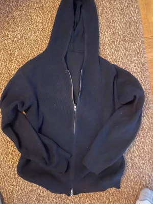 Svart stickad hoodie med dragkedja merinoull  - En svart stickad hoodie med hel dragkedja framtill och stor huva. Tröjan har ribbad struktur och långa ärmar, perfekt för en avslappnad och stilren look. Passar dig som gillar enkel och klassisk streetwear.